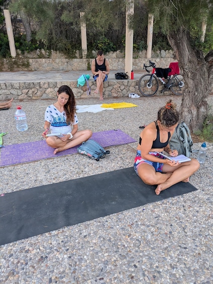 Two people meditating on mats outdoors with a tree and a stone wall in the background. 4