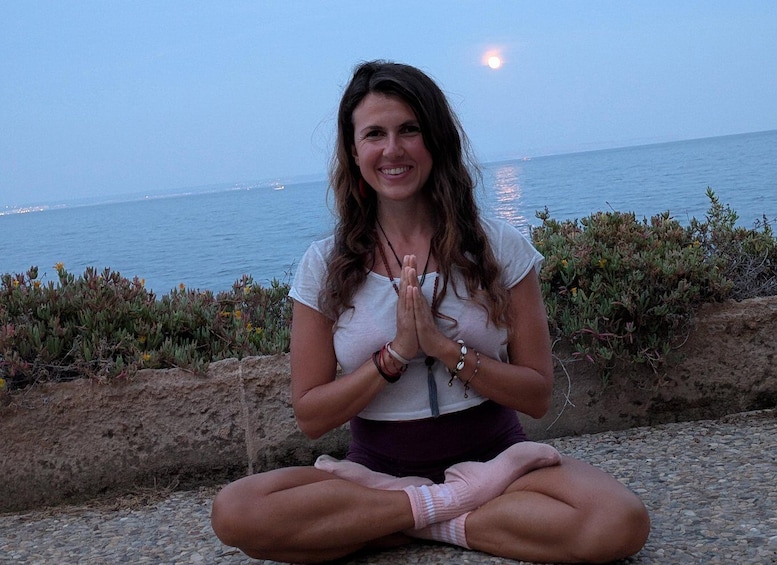 A person sitting cross-legged on a stone surface by the sea, with a rocky shoreline and ocean in the background. 2