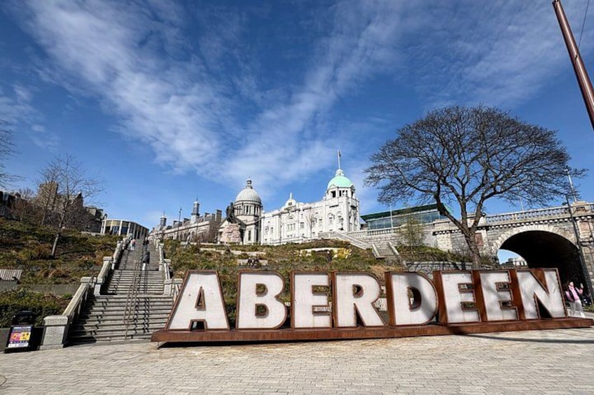 A large sign with the word 'ABERDEEN' in the foreground, a staircase leading up to a building with a dome, and a bridge in the background. 1