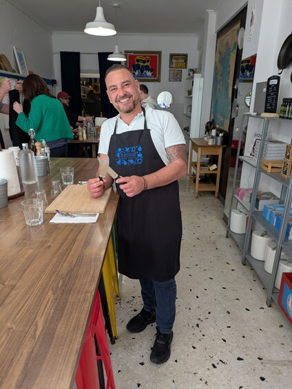 A person in a kitchen holding a knife and a cutting board, with various kitchen items and a map on the wall in the background. 1