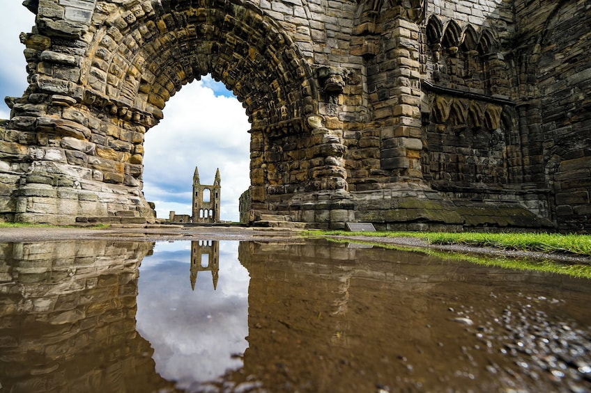 An ancient stone archway with a reflection in the water, leading to a ruined structure with two towers. 1