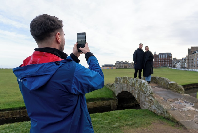 A person taking a photo with a smartphone, a stone bridge, and a grassy field with buildings in the background. 3