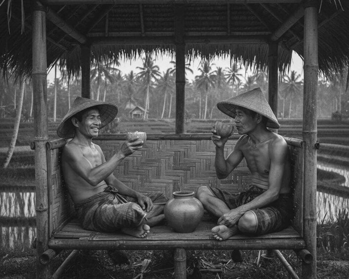 Two people sitting on a wooden bench in a rural setting, wearing conical hats, with a thatched roof structure and palm trees in the background. 2