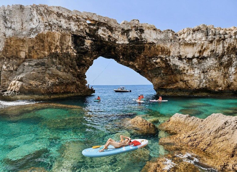A rocky arch formation with clear turquoise water, people floating on rafts, and a boat in the distance. 1