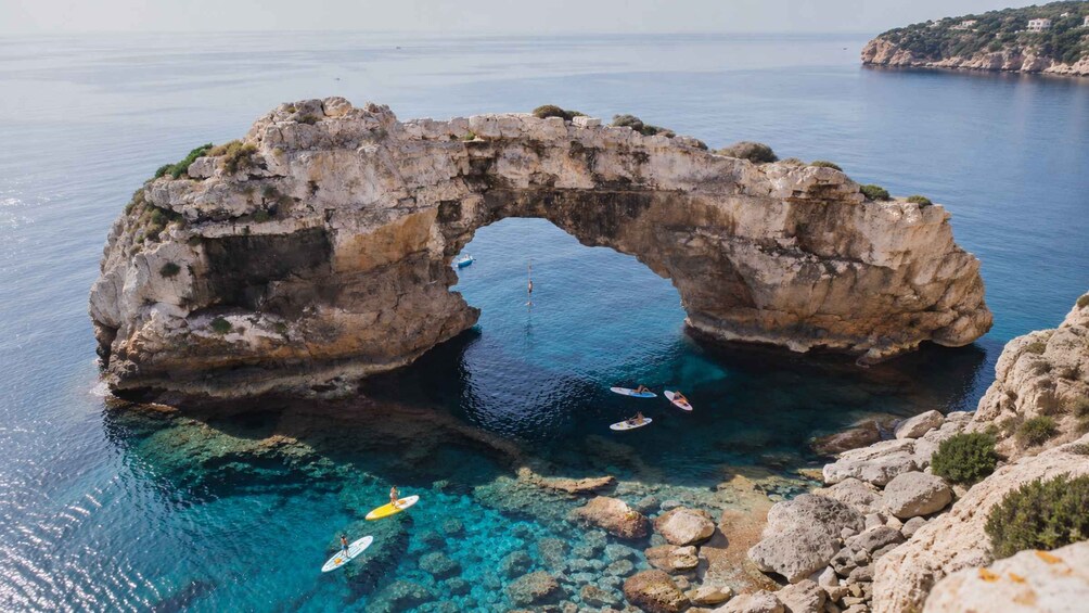 A natural rock arch formation in clear blue waters with people surfing. 1
