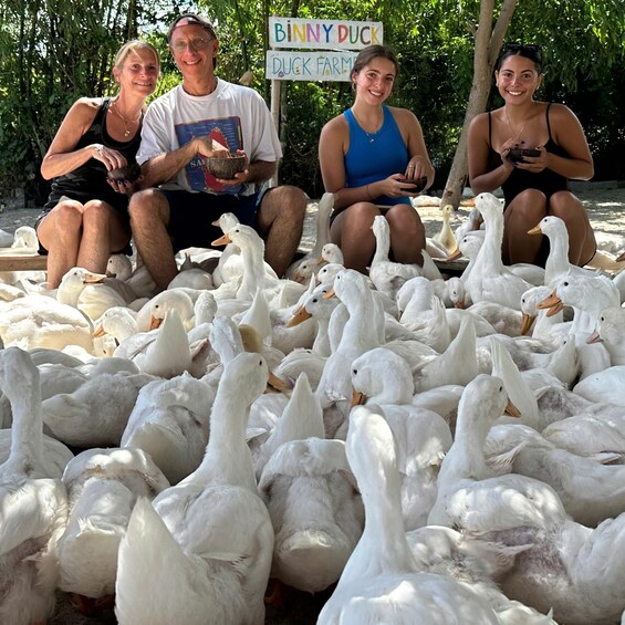 A group of people sitting among a large flock of white ducks. 4