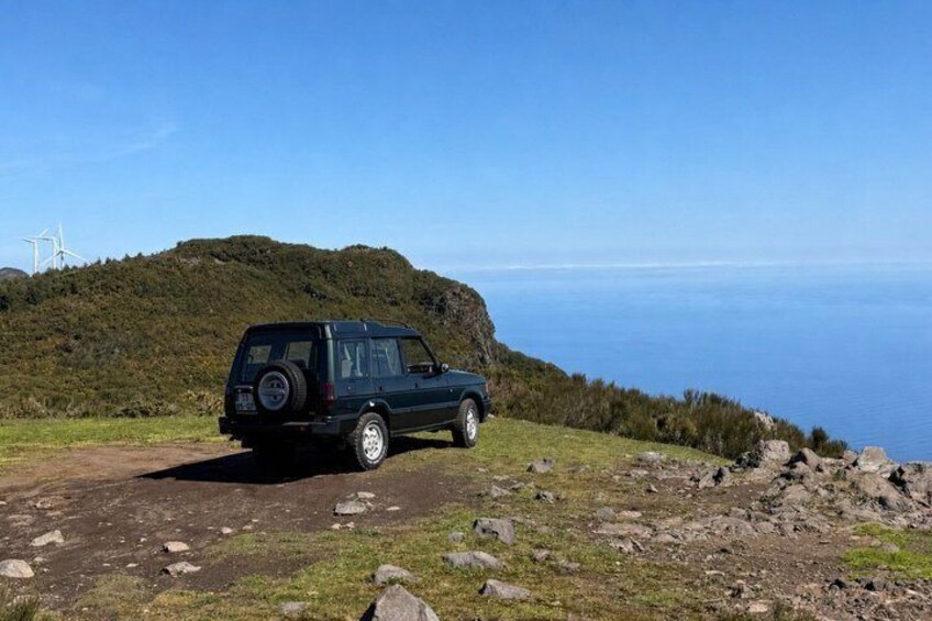 A dark-colored SUV parked on a dirt path with a coastal view and wind turbines in the background. 2