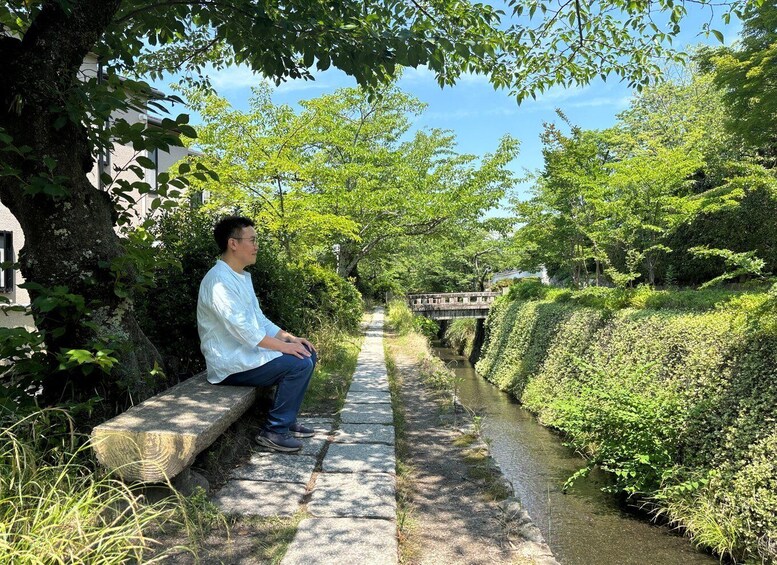 A man sitting on a stone bench by a narrow waterway, with lush greenery and a bridge in the background. 5