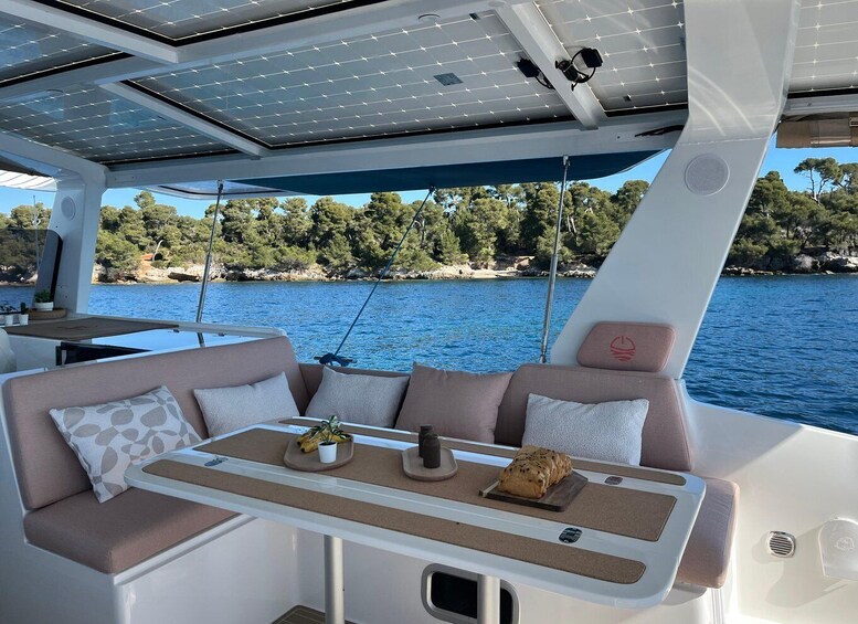 A boat interior with a cushioned seating area, a table with a plant and a loaf of bread, and a view of a calm lake with trees and rocks in the background. 5