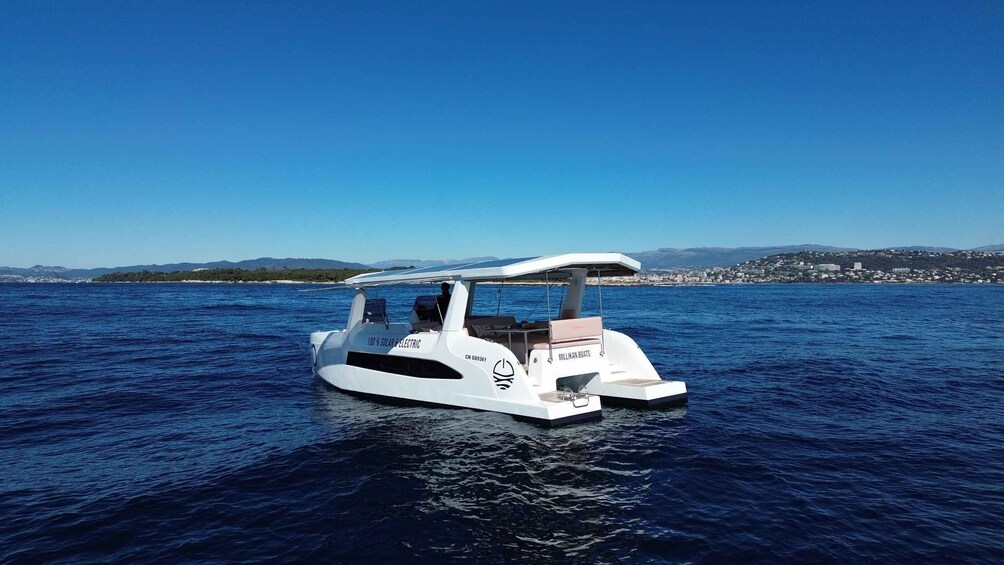 A white electric boat named '100% SOLAR & ELECTRIC' on calm waters with a clear blue sky and distant land in the background. 1