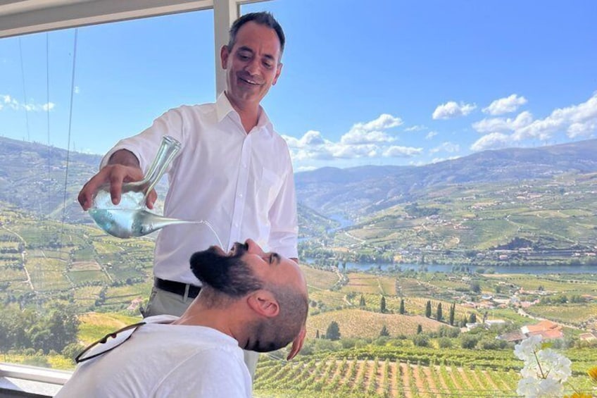 A person pouring a drink from a glass into another's mouth, with a scenic vineyard and hills in the background. 5
