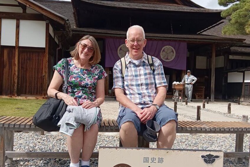 Two people sitting on a bench in front of a traditional Japanese building with a purple banner. 4