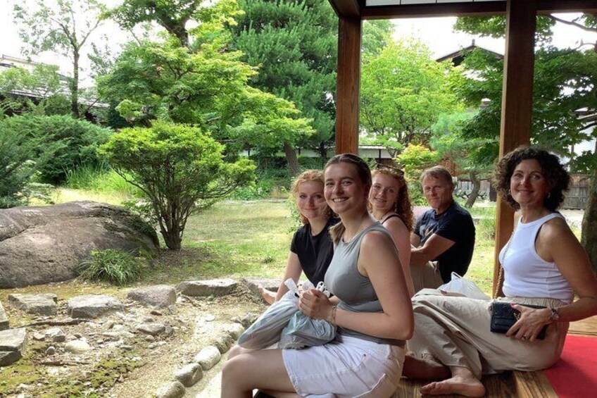 A group of people sitting on a wooden bench in a garden with rocks and trees. 2