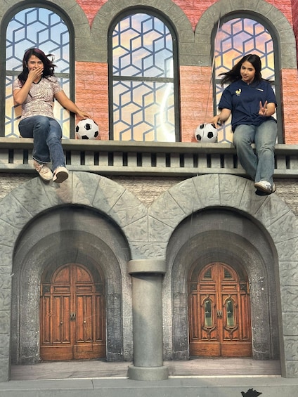 Two individuals sitting on a stone ledge with a soccer ball, overlooking a building with arched windows and wooden doors. 3