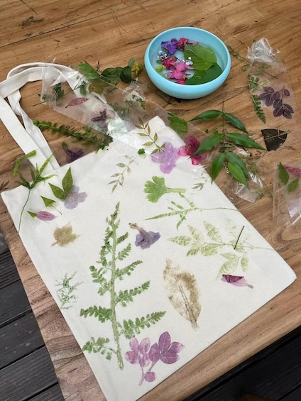 A fabric tote bag with embroidered plants and flowers, a blue bowl with water and leaves, and plastic bags with dried plants on a wooden surface. 1