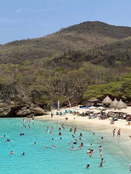 A beach with clear turquoise water, people swimming and sunbathing, and a mountainous backdrop. 4