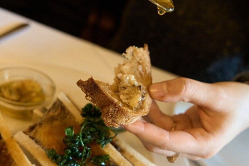 A hand holding a piece of bread with a filling, a small bowl of sauce, and a sprig of parsley on a table. 1