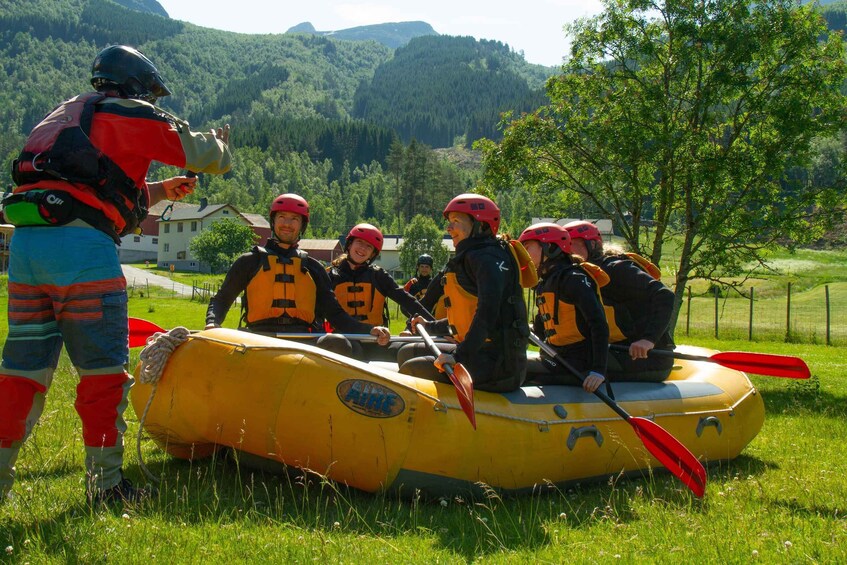 A group of people in a yellow raft with red paddles, wearing helmets and life jackets, on a grassy field with trees and mountains in the background. 5