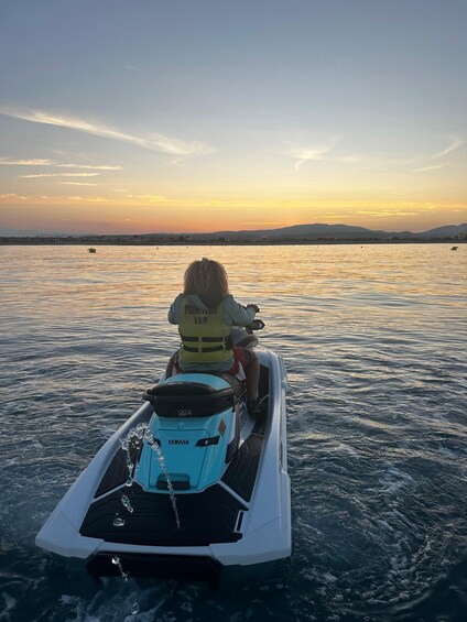 A person on a jet ski with a life jacket, heading towards a sunset over calm water. 1