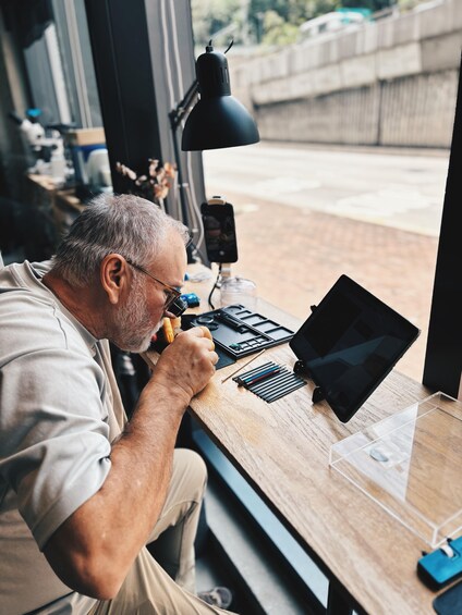 A man working at a desk with tools and a laptop, near a window overlooking a street. 3