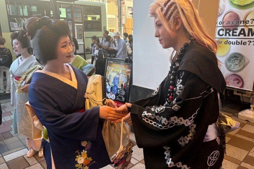 Two individuals in traditional attire, one in a blue kimono and the other in a black outfit with chains, standing in a public indoor space with a poster in the background. 4