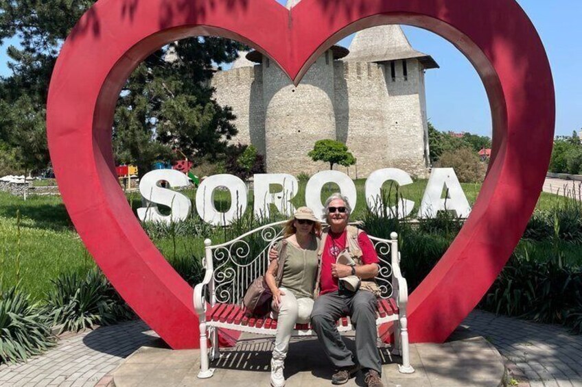A heart-shaped sign with 'SORGICA' in the center, a bench, and a stone building in the background. 2