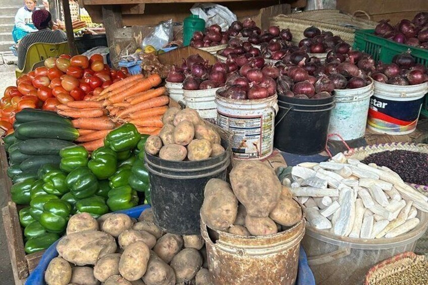 A market stall with various vegetables and fruits, including tomatoes, carrots, green peppers, potatoes, onions, and a container of fish. 2
