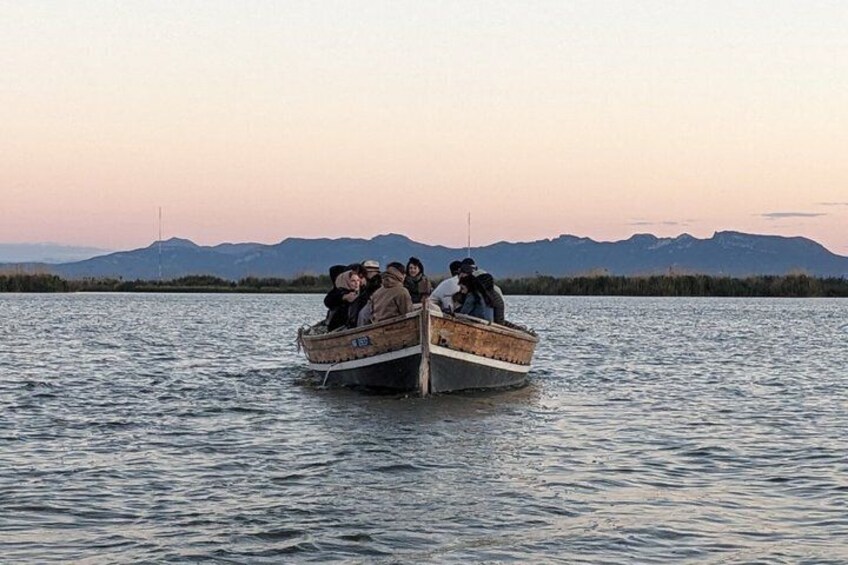 A group of people in a small wooden boat on a calm body of water with mountains in the background. 2