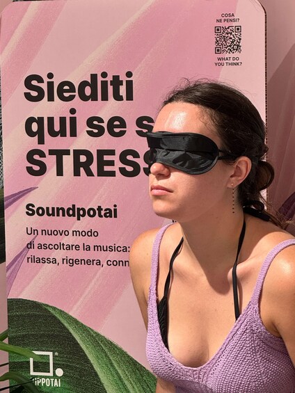 A person with a blindfold, wearing a purple top, standing in front of a pink background with text and a QR code. 1