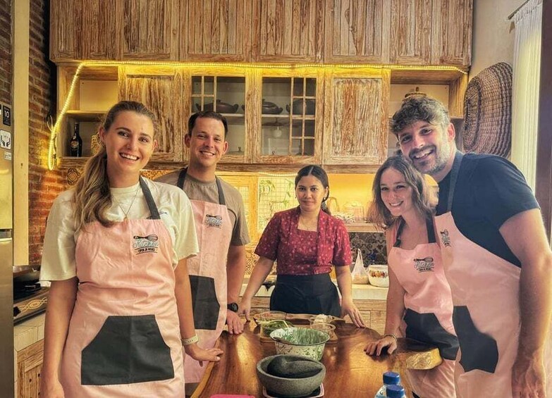 A group of people in aprons standing in a kitchen with wooden cabinets and a brick wall. 1
