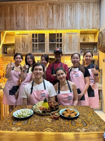 A group of people in aprons standing in a kitchen with wooden cabinets and a patterned tablecloth. 5