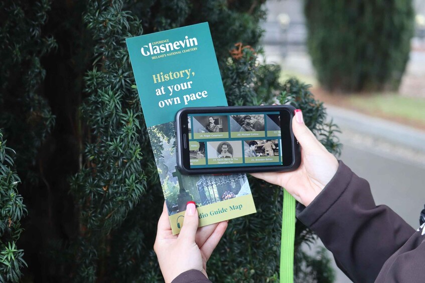 A person holding a book titled 'Experience Glasnevin' and a smartphone with a guide map. 1