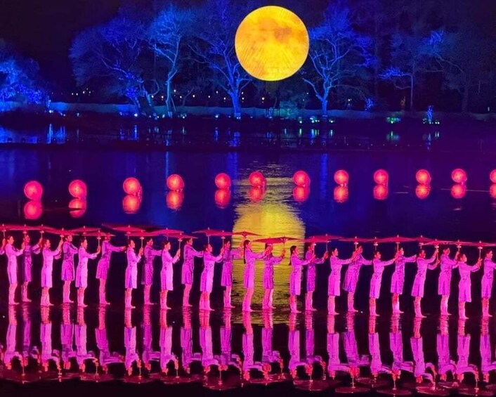 A nighttime outdoor scene with a large illuminated full moon, a reflective water surface, and a group of people in pink costumes standing in a line. 3