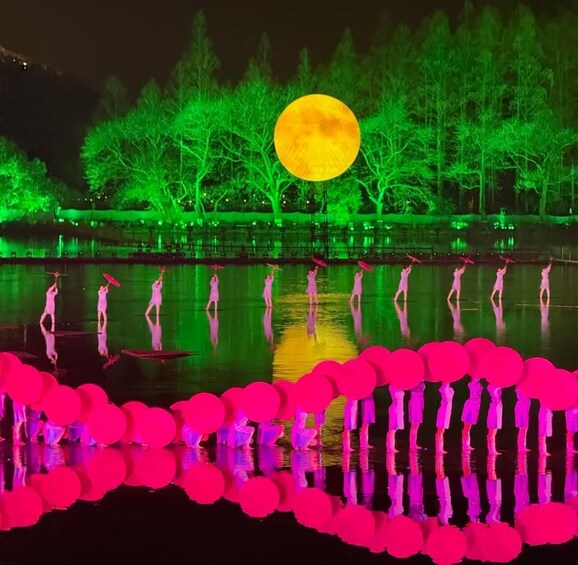 A nighttime outdoor scene with performers on stilts, a large illuminated moon, and a body of water reflecting the scene. 5