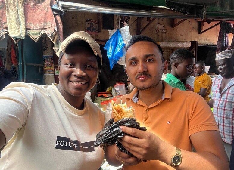 Two people holding food items in a market setting with other individuals in the background. 5