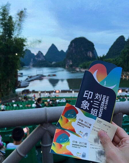 A person holding a colorful brochure with text in Chinese, a scenic view of a river with mountains in the background, and a crowd of people on a bridge. 4