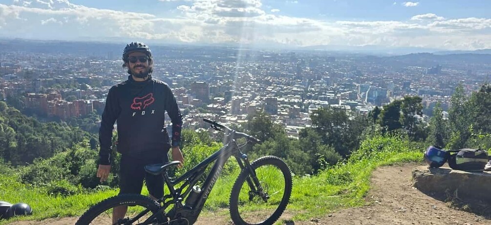 A person standing next to a mountain bike on a trail with a cityscape in the background. 4