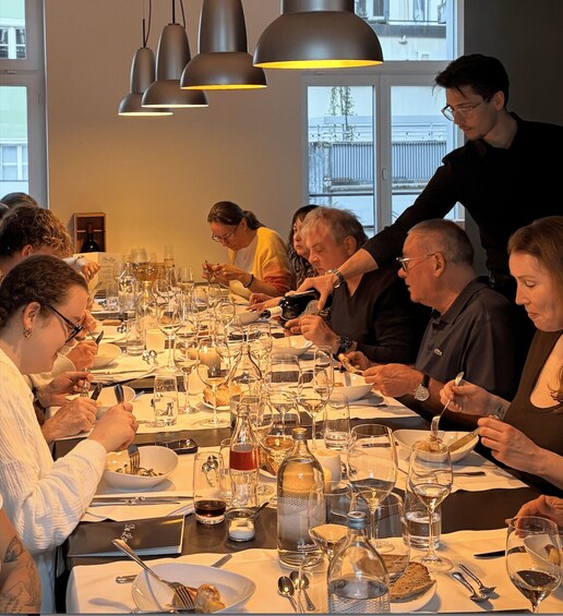 A group of people dining at a long table with wine glasses, bottles, and plates. 3