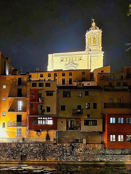 A night view of a coastal town with a prominent church tower, colorful buildings, and a stone wall by the water. 1