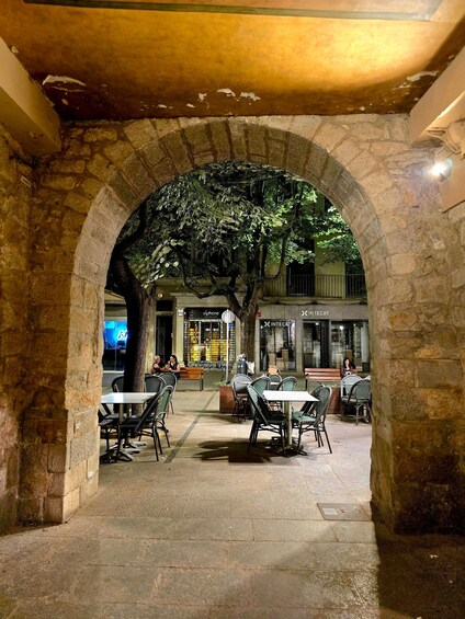 An outdoor café with stone arches, tables, and chairs, under a tree. 2