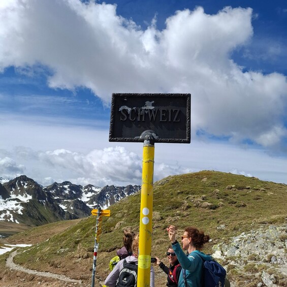 A signpost with 'SCHWEIZ' indicating a location, hikers on a mountain trail, and snow-capped peaks in the background. 3