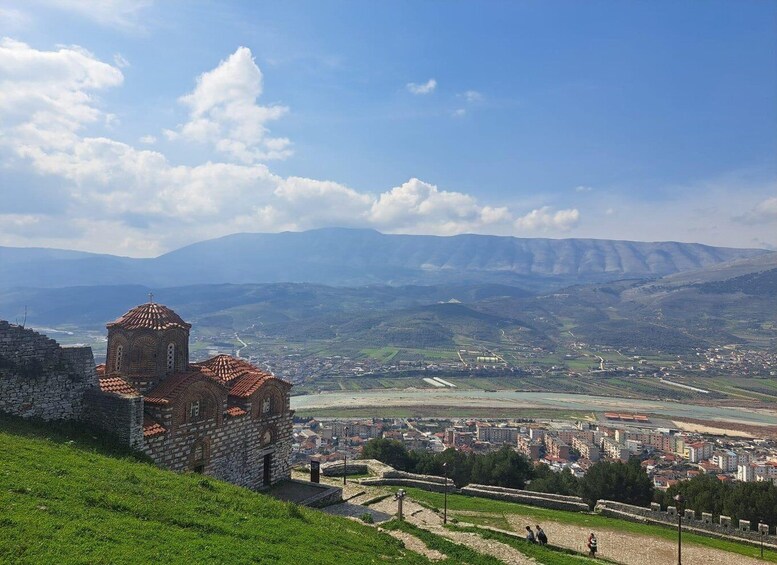 A historic stone church with a red-tiled roof, a cityscape with buildings and a river, and rolling hills in the background. 1