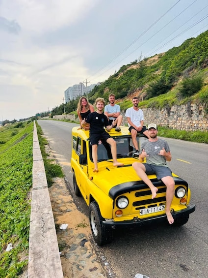 A yellow vehicle with four people on the roof, parked on a roadside with greenery and a stone wall. 1