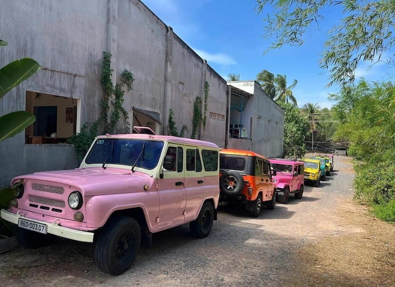 A row of vintage vehicles parked along a dirt path beside a concrete building. 3