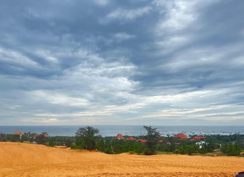 A coastal landscape with sandy dunes, a beach, and a town with red-roofed buildings. 5