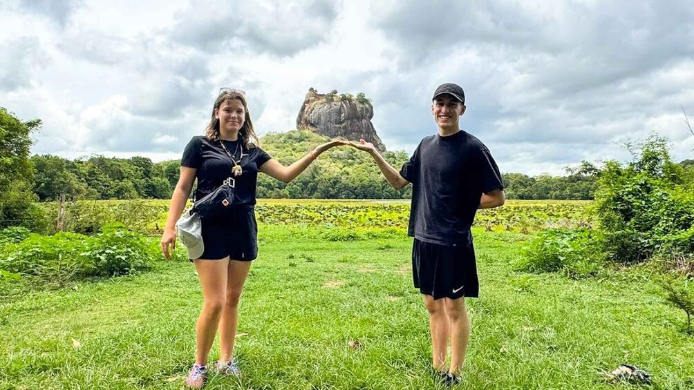 Two people holding a rock formation in a grassy field with trees and a cloudy sky. 1