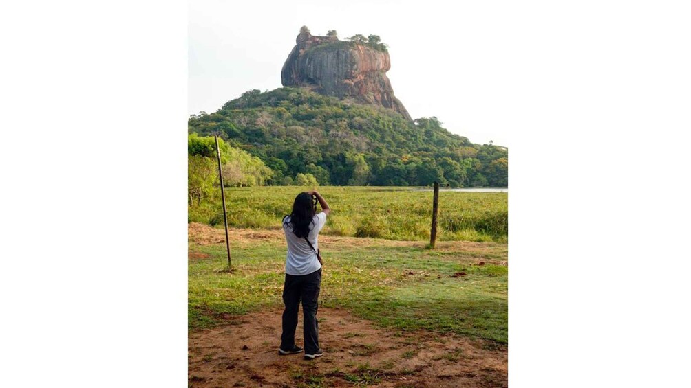 A person taking a photo of a large rock formation surrounded by greenery. 5