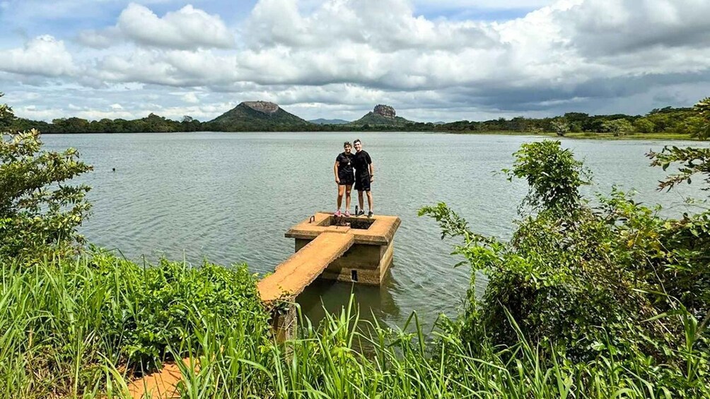 Two people standing on a concrete platform extending into a lake, surrounded by greenery and hills in the background. 3