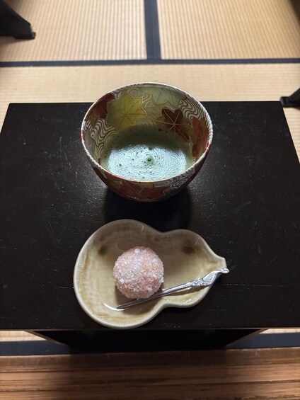 A bowl of green tea with a decorative pattern, a small dish with a pink sugar-coated item, and a silver spoon on a black table. 3