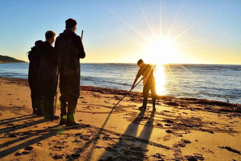 Three people on a beach at sunset, one person is using a stick to collect seaweed. 3